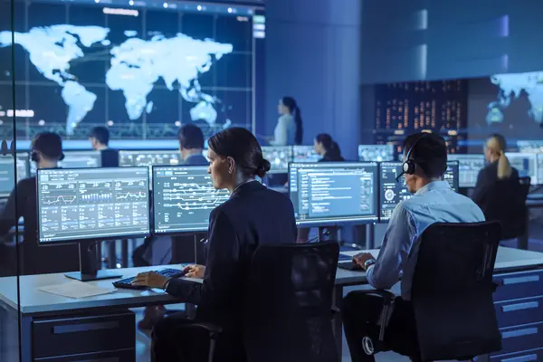 Moderner Kontrollraum mit mehreren Personen an Computerarbeitsplätzen, die Daten und Aktivitäten auf Bildschirmen überwachen. Im Hintergrund ist eine große Weltkarte auf einem Bildschirm zu sehen.