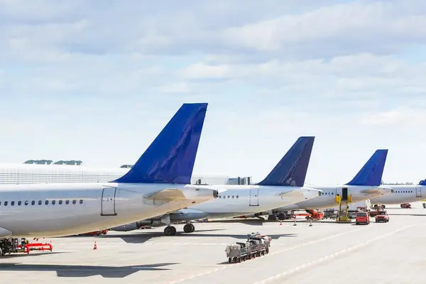 Reihe von Verkehrsflugzeugen, die auf dem Vorfeld eines Flughafens geparkt sind, mit sichtbaren Heckflossen und im Einsatz befindlichen Bodenfahrzeugen.