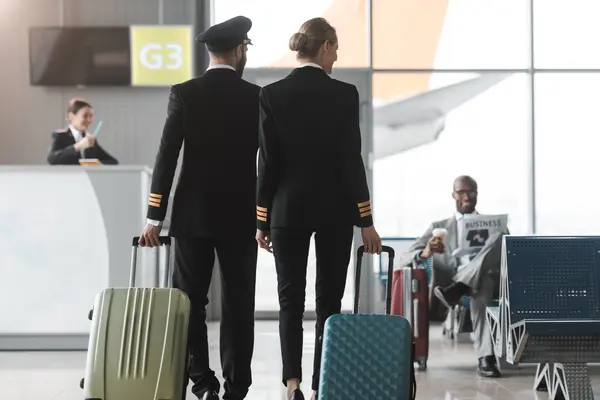 Two pilots in uniforms walking with rolling suitcases through an airport, with an airplane and a waiting passenger in the background.