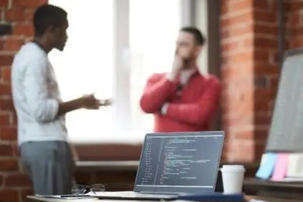 Laptop displaying programming code on a desk, two people talking in the background.