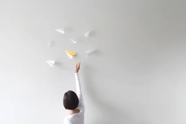 Person reaching for a yellow paper airplane surrounded by white paper airplanes against a bright wall.