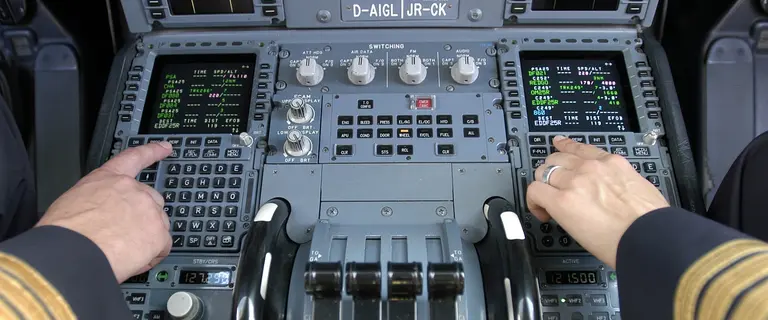 Cockpit eines Flugzeugs mit Fokus auf das zentrale Bedienfeld und zwei Flight-Management-System-Displays (FMS), die Flugdaten wie Wegpunkte, Geschwindigkeit und Flughöhe anzeigen. Zwei Hände bedienen die Steuerungselemente.