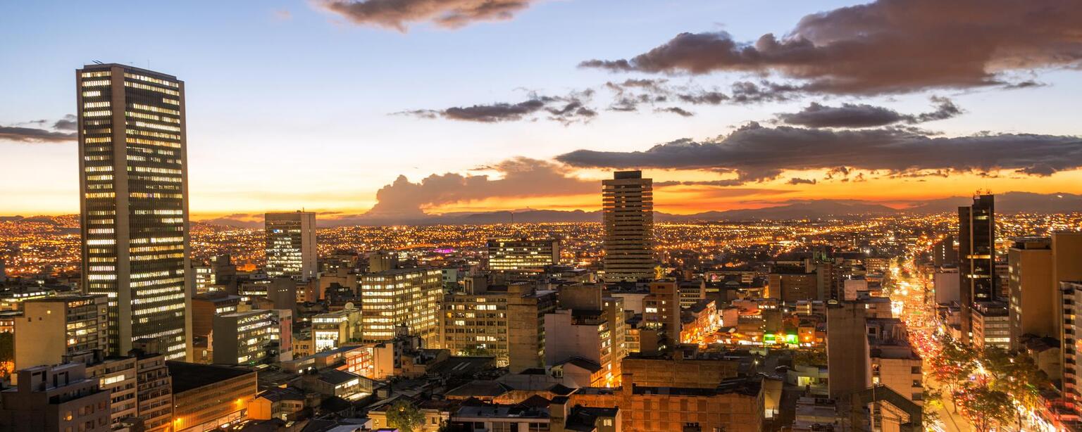 A panoramic aerial view of Bogota, Colombia, as the sun sets. The city is bathed in a warm glow of orange and pink, and the skyscrapers stand tall against the darkening sky.