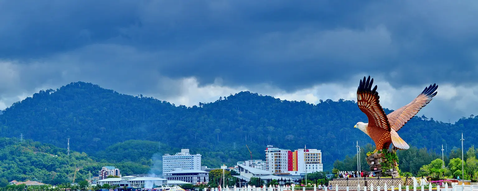 A large statue of a bird, possibly an eagle or hawk, with its wings outstretched, flying over a body of water in front of a city skyline. The statue is located in Langkawi, Malaysia, and is known as Eagle Square.