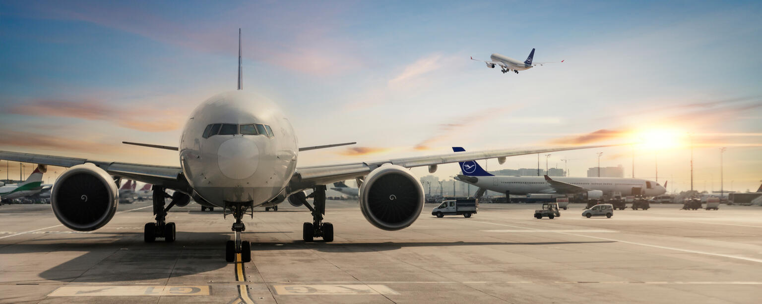 A large commercial airplane is positioned on the tarmac at an airport, facing the viewer. The aircraft's engines and landing gear are visible. In the background, another airplane is taking off, and multiple other aircraft and airport ground vehicles are present. The scene is set against a sunrise or sunset sky with warm lighting.